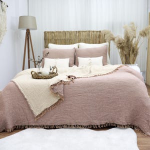 May include: A bed with a woven headboard, covered in a dusty rose-colored quilt and pillows. A cream-colored throw with fringe is draped over the quilt. A woven tray with candles and a vase of greenery sits on the bed. A wooden floor lamp stands to the left.