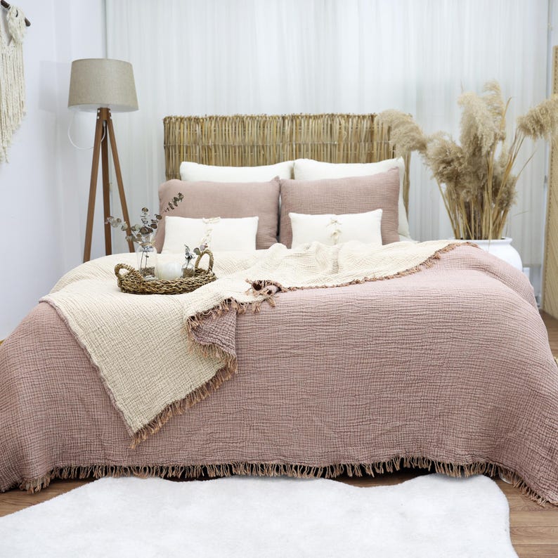 May include: A bed with a woven headboard, covered in a dusty rose-colored quilt and pillows. A cream-colored throw with fringe is draped over the quilt. A woven tray with candles and a vase of greenery sits on the bed. A wooden floor lamp stands to the left.