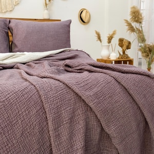 May include: A soft, purple, textured throw blanket with fringe edges is draped over a bed with a white and purple pillow. The bed is made with white sheets and a purple pillow.