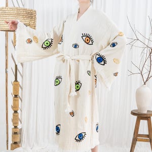 Pode incluir: Robe creme com estampa de olhos em v&aacute;rias cores. O robe tem mangas largas, cinto para amarrar e bainha com franjas. Os desenhos dos olhos apresentam detalhes azuis, verdes, laranja e amarelos. O robe &eacute; feito de tecido texturizado.