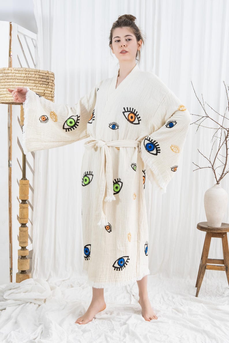Pode incluir: Robe creme com estampa de olhos em v&aacute;rias cores. O robe tem mangas largas, cinto para amarrar e bainha com franjas. Os desenhos dos olhos apresentam detalhes azuis, verdes, laranja e amarelos. O robe &eacute; feito de tecido texturizado.