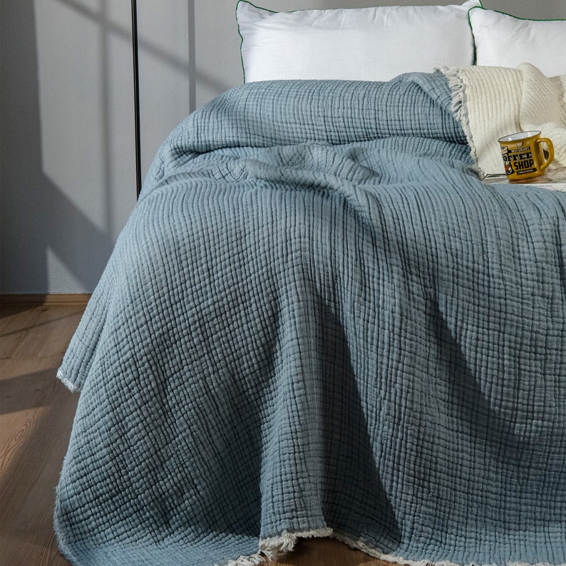 K&ouml;nnte beinhalten: Eine hellblaue, strukturierte Tagesdecke mit Fransenrand. Die Decke ist gefaltet und &uuml;ber ein Bett mit wei&szlig;en Kissen gelegt.