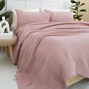 May include: A bed with a pink waffle-weave bedspread and matching pillowcases. The bed is made up with a blanket and two pillows. The bed is in a bedroom with a white headboard and a small side table.