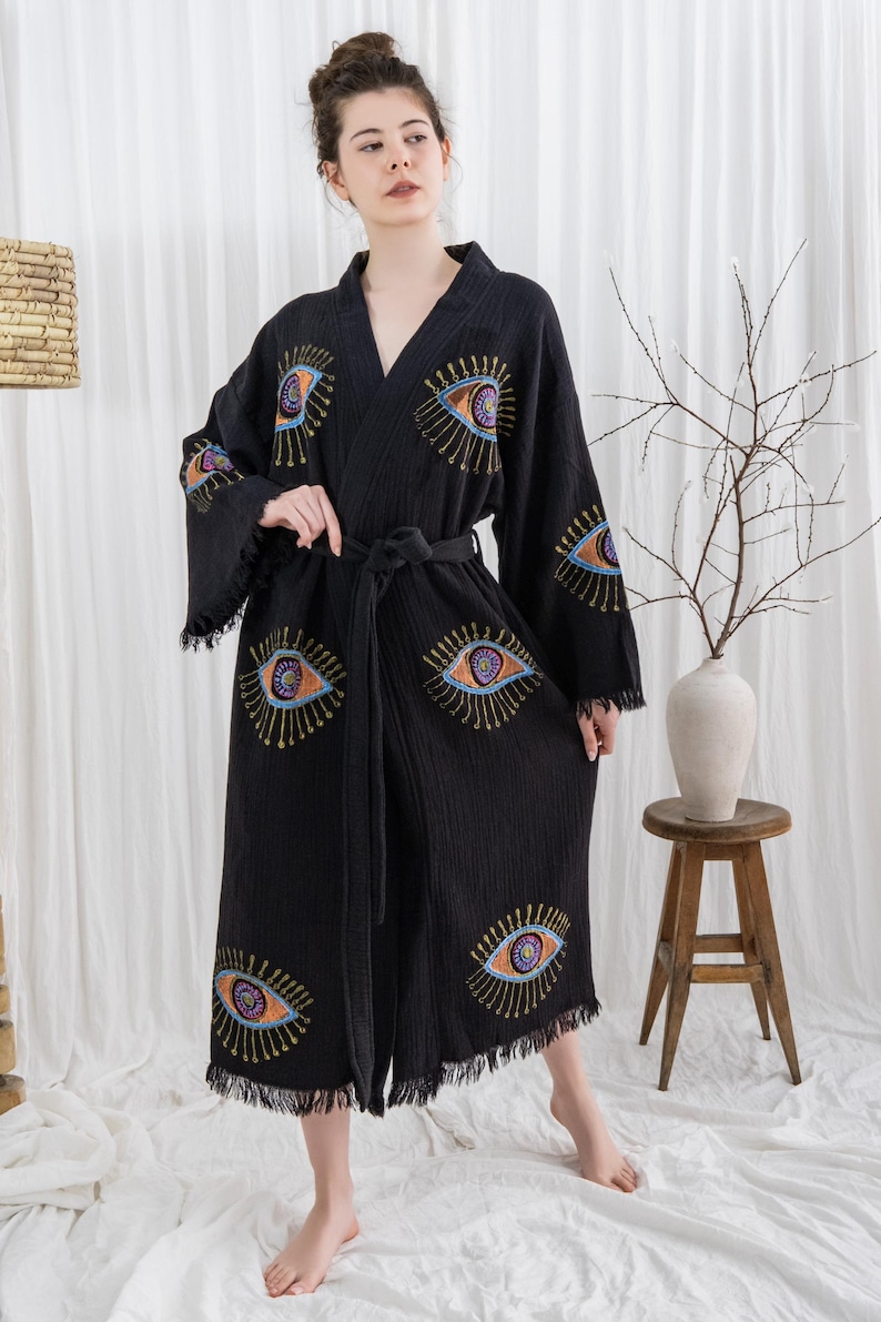 Pode incluir: Robe preta com desenhos de olhos bordados em laranja, azul e roxo. O robe tem uma bainha com franjas e um cinto a condizer. As mangas s&atilde;o compridas e largas, e o robe &eacute; feito de um tecido macio e texturizado.