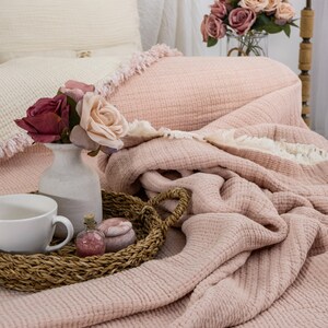 May include: A soft pink waffle weave throw blanket with a white fringe border. The blanket is folded and draped over a bed with a white pillow and a vase of pink roses.