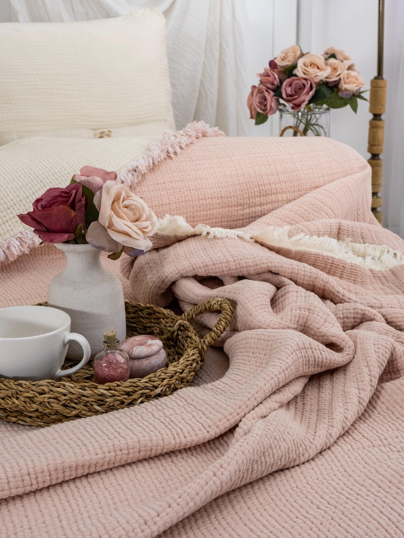 May include: A soft pink waffle weave throw blanket with a white fringe border. The blanket is folded and draped over a bed with a white pillow and a vase of pink roses.