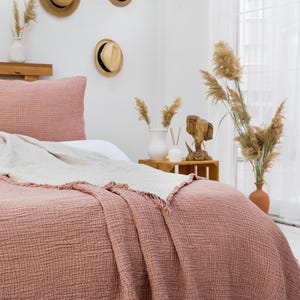 Puede incluir: Una manta de tejido de gofre rosa claro con bordes deshilachados est&aacute; colocada sobre una cama con una almohada blanca y una almohada rosa claro. La cama est&aacute; en un dormitorio con una pared blanca y una mesita de noche de madera con un jarr&oacute;n de flores secas.