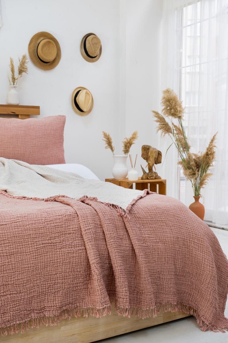 Puede incluir: Una manta de tejido de gofre rosa claro con bordes deshilachados est&aacute; colocada sobre una cama con una almohada blanca y una almohada rosa claro. La cama est&aacute; en un dormitorio con una pared blanca y una mesita de noche de madera con un jarr&oacute;n de flores secas.