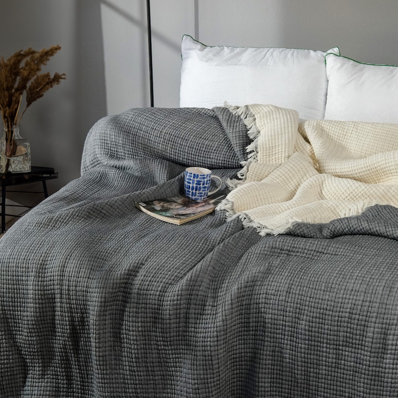 K&ouml;nnte beinhalten: Eine graue Waffel-Decke mit einer wei&szlig;en Waffel-Decke darauf. Die Decken liegen auf einem Bett mit wei&szlig;en Kissen. Eine blaue und wei&szlig;e Keramiktasse und ein Buch liegen auf den Decken.