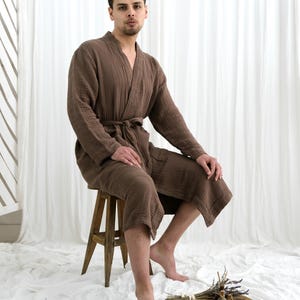 May include: A brown, knee-length robe with a tie waist. The robe is made of a textured fabric and is worn by a person sitting on a wooden stool. The background is a white curtain.