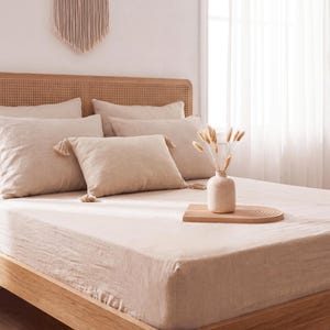 Può includere: Una struttura letto in legno chiaro con testiera in paglia di Vienna, vestita con biancheria da letto in lino beige che include due grandi cuscini quadrati, due cuscini rettangolari con nappe agli angoli e un lenzuolo con angoli. Un vaso beige con steli secchi poggia su un vassoio di legno.