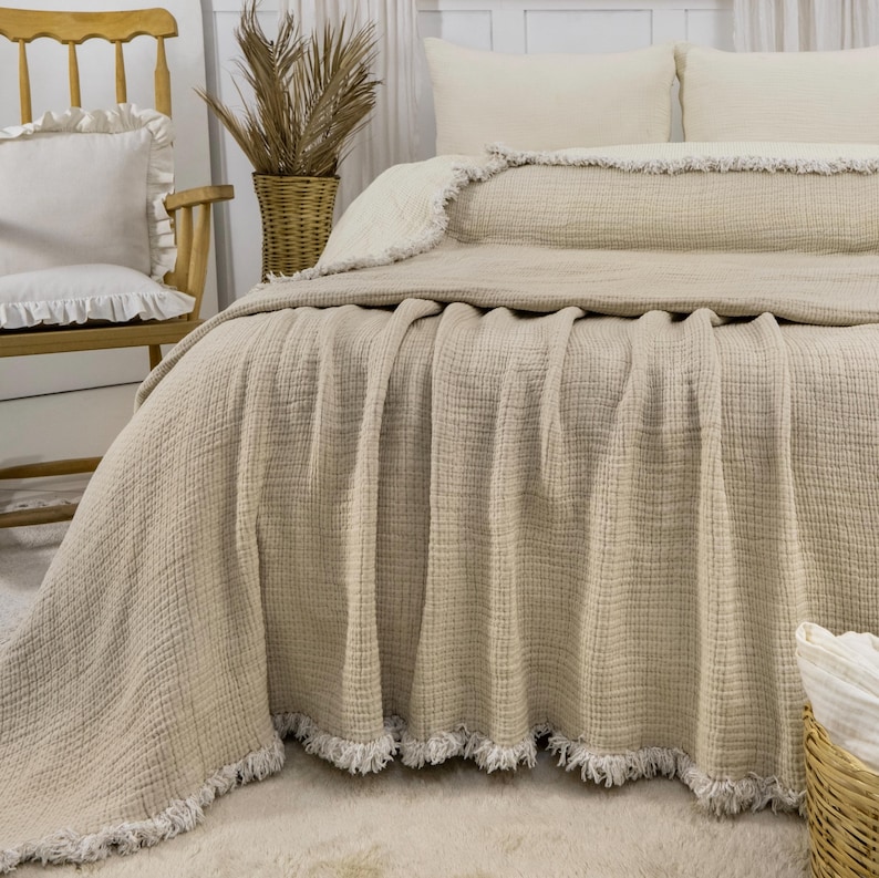 May include: A beige waffle weave throw blanket with a white fringe border. The blanket is folded on a bed with white pillows and a wooden chair in the background.