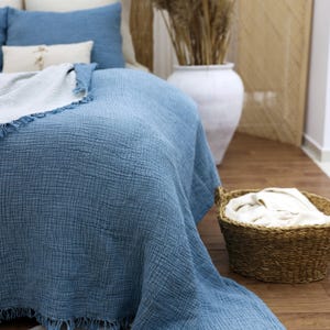 May include: A blue bedspread with a textured weave covers a bed, complemented by matching pillows. A white throw with fringe rests on the bed. A woven basket holds folded white linens. A large white vase with dried plants is in the background.