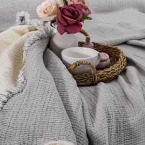 May include: A gray and white throw blanket with a fringe border. A white ceramic vase with pink and white roses sits on top of the blanket. A white mug and a wicker basket with a pink and white decorative item are also on the blanket.