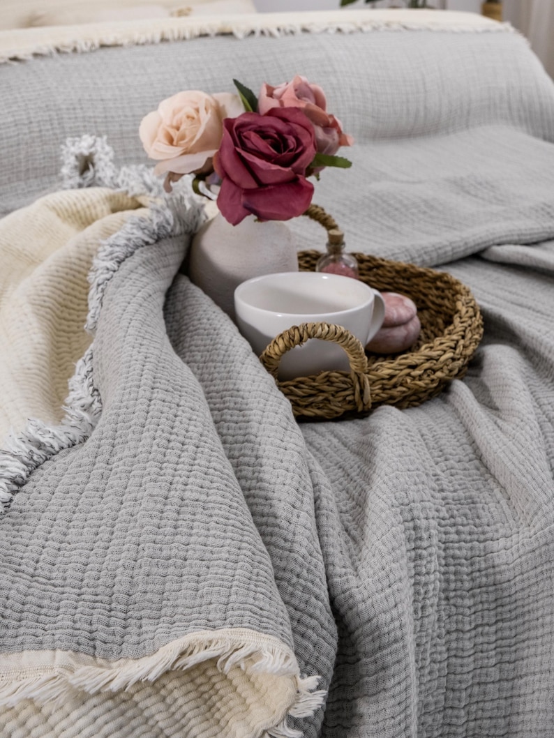 May include: A gray and white throw blanket with a fringe border. A white ceramic vase with pink and white roses sits on top of the blanket. A white mug and a wicker basket with a pink and white decorative item are also on the blanket.