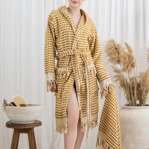 May include: A mustard yellow and white checkered hooded bathrobe with a matching towel. The robe has a belt, pockets, and fringe detailing. A wooden stool holds a bowl with bath accessories.