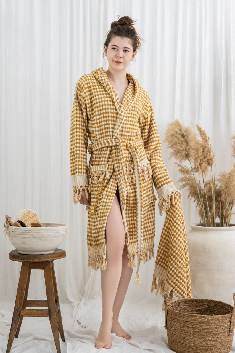 May include: A mustard yellow and white checkered hooded bathrobe with a matching towel. The robe has a belt, pockets, and fringe detailing. A wooden stool holds a bowl with bath accessories.