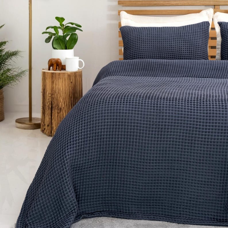 May include: A navy blue waffle-weave bedspread and matching pillow on a bed with a wooden headboard. The bed is made up with a white pillow and a navy blue pillow. A small wooden side table holds a plant, a mug, and a small elephant figurine.