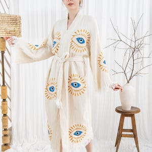Pode incluir: Robe creme com um desenho de olhos repetido em azul e dourado. O robe tem uma cintura com la&ccedil;o e bordas com franjas. As mangas s&atilde;o largas e esvoa&ccedil;antes. O robe &eacute; feito de um tecido texturizado.