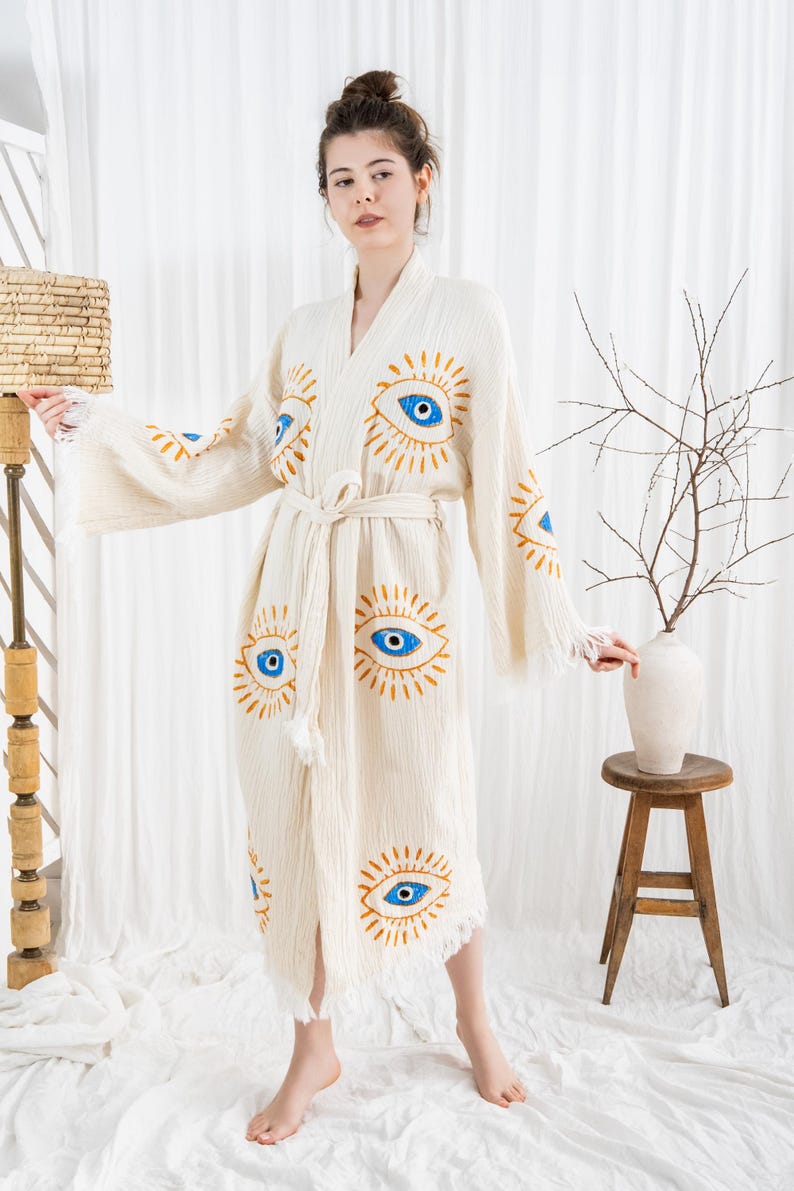 Pode incluir: Robe creme com um desenho de olhos repetido em azul e dourado. O robe tem uma cintura com la&ccedil;o e bordas com franjas. As mangas s&atilde;o largas e esvoa&ccedil;antes. O robe &eacute; feito de um tecido texturizado.