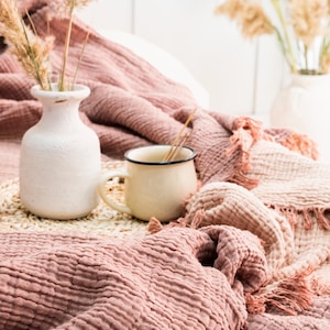 Pode incluir: Um close-up de um cobertor texturizado rosa e branco com um vaso de cerâmica branco e uma caneca de esmalte branco em um tapete tecido.