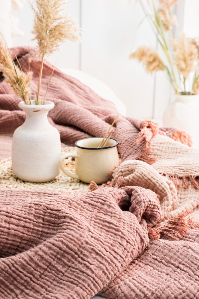 Puede incluir: Primer plano de una manta de textura rosa y blanca con un jarr&oacute;n de cer&aacute;mica blanco y una taza de esmalte blanco sobre una estera tejida.