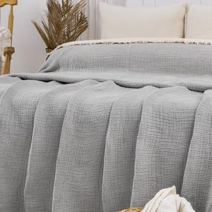 May include: A gray waffle weave throw blanket with a fringe border. The blanket is folded on a bed with white pillows and a wicker basket with white towels.