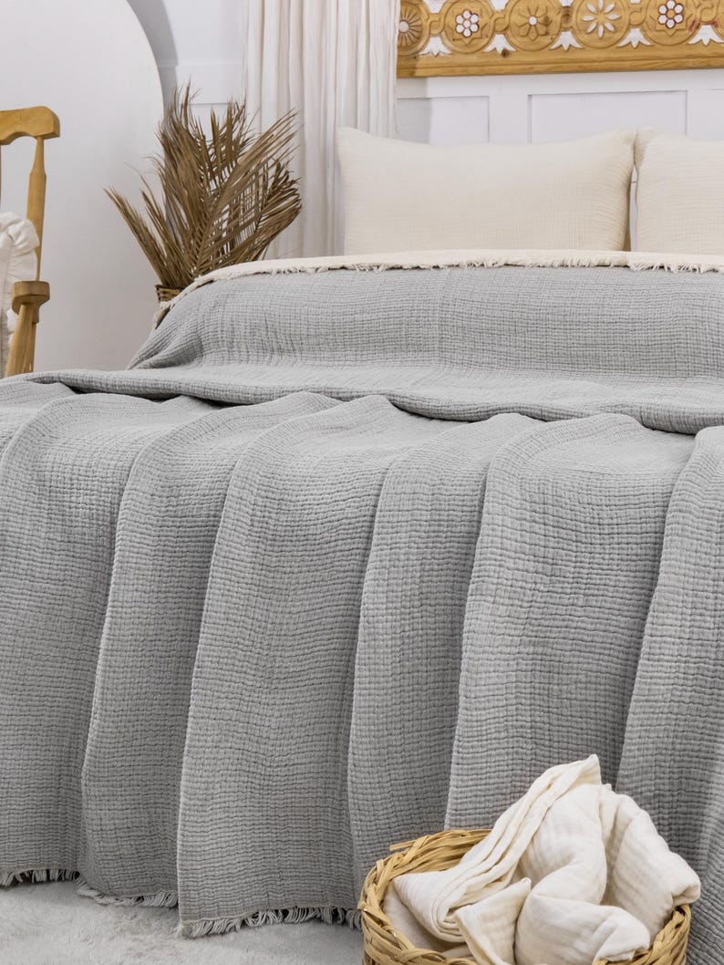 May include: A gray waffle weave throw blanket with a fringe border. The blanket is folded on a bed with white pillows and a wicker basket with white towels.