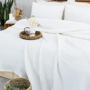 May include: A white waffle-weave bedspread and pillow set. A woven tray with a white ceramic pitcher and flowers sits on the bed. The bed is made up with white sheets and pillows. A woven basket with greenery is in the foreground.