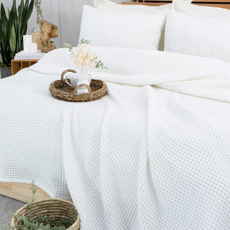 May include: A white waffle-weave bedspread and pillow set. A woven tray with a white ceramic pitcher and flowers sits on the bed. The bed is made up with white sheets and pillows. A woven basket with greenery is in the foreground.