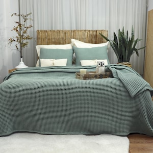 May include: A bed is made with a sage green waffle-weave blanket and matching pillows. The headboard is made of woven natural material. A wooden tray with a candle and owl figurine sits on the bed. A white rug is on the floor.