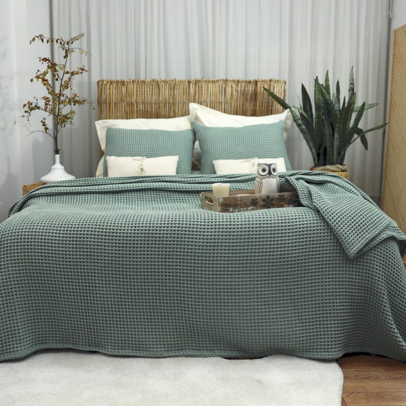 May include: A bed is made with a sage green waffle-weave blanket and matching pillows. The headboard is made of woven natural material. A wooden tray with a candle and owl figurine sits on the bed. A white rug is on the floor.
