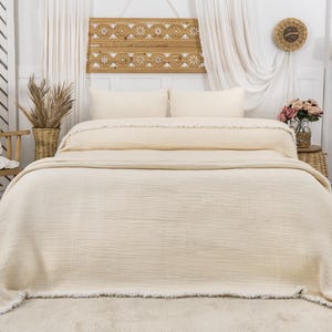 May include: A cream-colored waffle weave throw blanket with a fringe border. The blanket is folded on a bed with white pillows and a wooden headboard.