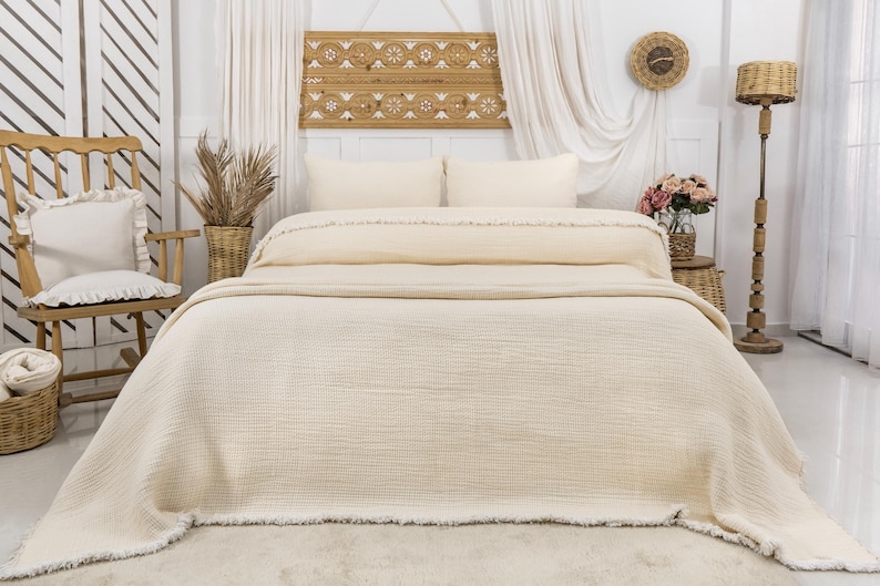 May include: A cream-colored waffle weave throw blanket with a fringe border. The blanket is folded on a bed with white pillows and a wooden headboard.