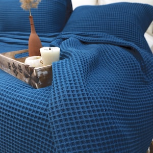 May include: A blue waffle-weave blanket draped over a bed with a matching pillow. A wooden tray holds candles and a small vase with dried flowers. The blanket's texture is visible, creating a cozy and inviting atmosphere.