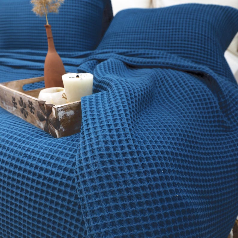 May include: A blue waffle-weave blanket draped over a bed with a matching pillow. A wooden tray holds candles and a small vase with dried flowers. The blanket's texture is visible, creating a cozy and inviting atmosphere.
