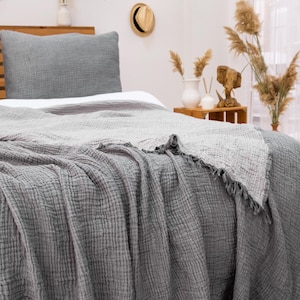 May include: A bed with a gray textured blanket and pillow. The blanket has a fringed edge. The bed is made up with a white sheet. Decorative hats hang on the wall above the bed. The room has a neutral color palette.
