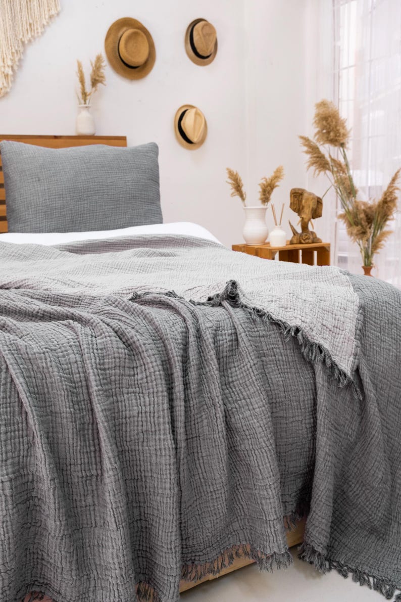 May include: A bed with a gray textured blanket and pillow. The blanket has a fringed edge. The bed is made up with a white sheet. Decorative hats hang on the wall above the bed. The room has a neutral color palette.