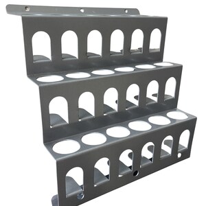 Könnte beinhalten: Ein grauer, mehrstufiger Metall-Organizer mit drei Ebenen, die jeweils bogenförmige Aussparungen und kreisförmige Öffnungen aufweisen. Die oberste Ebene hat eine flache Verlängerung mit Befestigungslöchern. Für zylindrische Gegenstände.