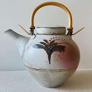 Op de afbeelding: Een ronde keramische theepot met een gespikkeld gebroken wit glazuur, voorzien van een handgeschilderd donkerbruin bloemmotief en een zachte roze kleurzweem. De pot heeft een gebogen tuit, een bijpassend deksel en een lichtgekleurd bamboe handvat.