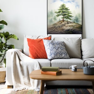 May include: A watercolor painting of a lone tree on a rocky cliff, surrounded by fog. The painting is framed in black and hangs on a white wall above a white couch with orange and blue patterned pillows.