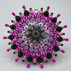Peut inclure: Un ornement décoratif rond composé d'un groupe central de cristaux gris foncé à facettes, entouré d'anneaux concentriques de sequins rose vif, de tissu à pois blancs et noirs, de perles de lave noires et de perles roses en forme de goutte.