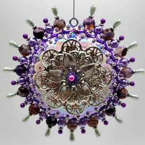 Puede incluir: Un adorno colgante redondo decorado con lentejuelas moradas e iridiscentes, cuentas de color morado oscuro y detalles claros en forma de lágrima. El centro presenta una flor de filigrana de metal plateado superpuesta con una pequeña cuenta rosa.