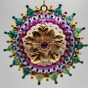 Peut inclure: Une décoration sphérique ornée d'une fleur centrale en métal doré avec un cœur violet, entourée de couches de sequins blancs, de sequins violets vifs et d'un périmètre de perles marbrées turquoise, de sphères dorées et de petites perles roses et bleues.