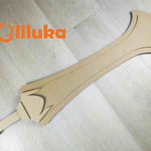 Puede incluir: Una espada grande de cartón sin terminar con un mango y una hoja decorativos. La espada es de color marrón claro y está sobre una superficie de madera gris claro. El logotipo "Oliluka" está en naranja en la esquina superior izquierda.