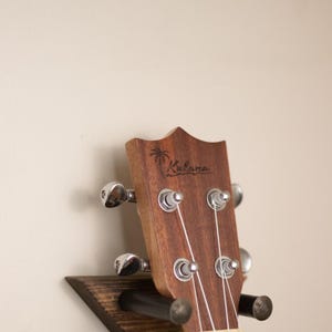 Może przedstawiać: Drewniany uchwyt ścienny na ukulele z czarnym metalowym hakiem. Na haku wisi brązowe ukulele z białymi strunami. Ukulele ma napis "Kala" na główce.