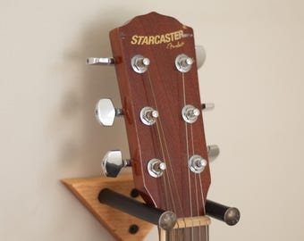 May include: A wooden guitar wall mount with a black metal bar, holding a brown acoustic guitar with the text "STARCASTER by Fender" on the headstock.