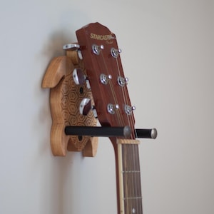 May include: A wooden turtle-shaped guitar wall mount with a black metal bar holding a guitar with a brown wooden body and a black and white fretboard. The guitar headstock has the text "STARCASTER" printed on it.