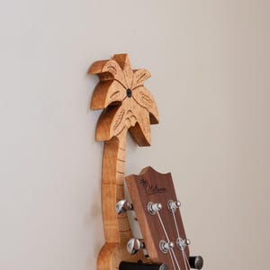Może przedstawiać: Drewniany uchwyt na ścianę do ukulele w kształcie palmy z wiszącym ukulele. Ukulele jest brązowe z białą struną, a na główce nadrukowana jest nazwa marki "Kala".
