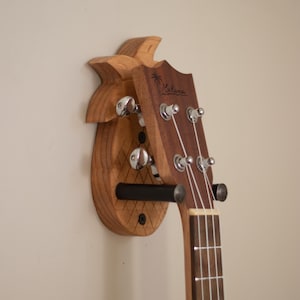 Może przedstawiać: Drewniany uchwyt ścienny do ukulele w kształcie ananasa z czarnym metalowym hakiem. Ukulele jest jasnobrązowe z białymi strunami i napisem "Kala Brand" na główce.